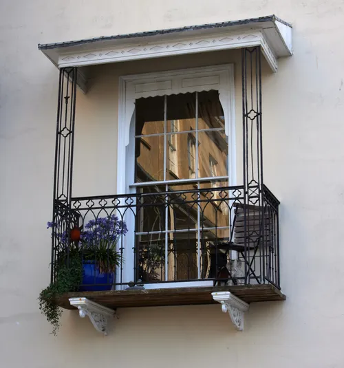 This architecture photograph taken in the morning during late summer shows a balcony and window on a residential building in Bristol, England, United Kingdom. The main subject is the balcony window, which is framed by wrought iron railings and decorated with flower pots and a wooden chair. The window features a notable reflection of adjacent buildings, demonstrating the typical urban style found in this part of Bristol. The image captures the soft morning light, highlighting the textures of the wall, the balcony structure, and the window’s intricate, reflective surface.